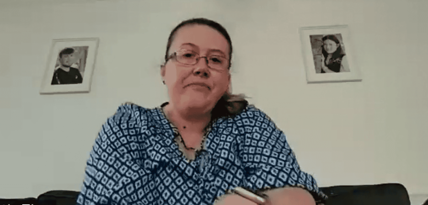 Woman in glasses blue top, photo frames behind.