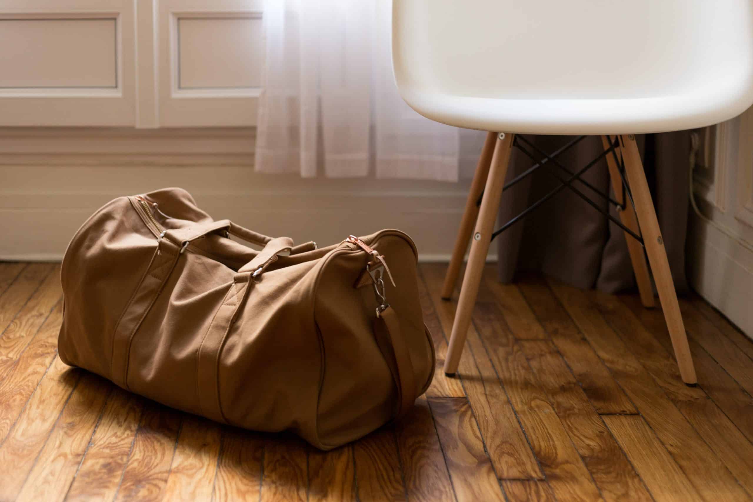 Brown duffel bag on wooden floor, white chair beside.